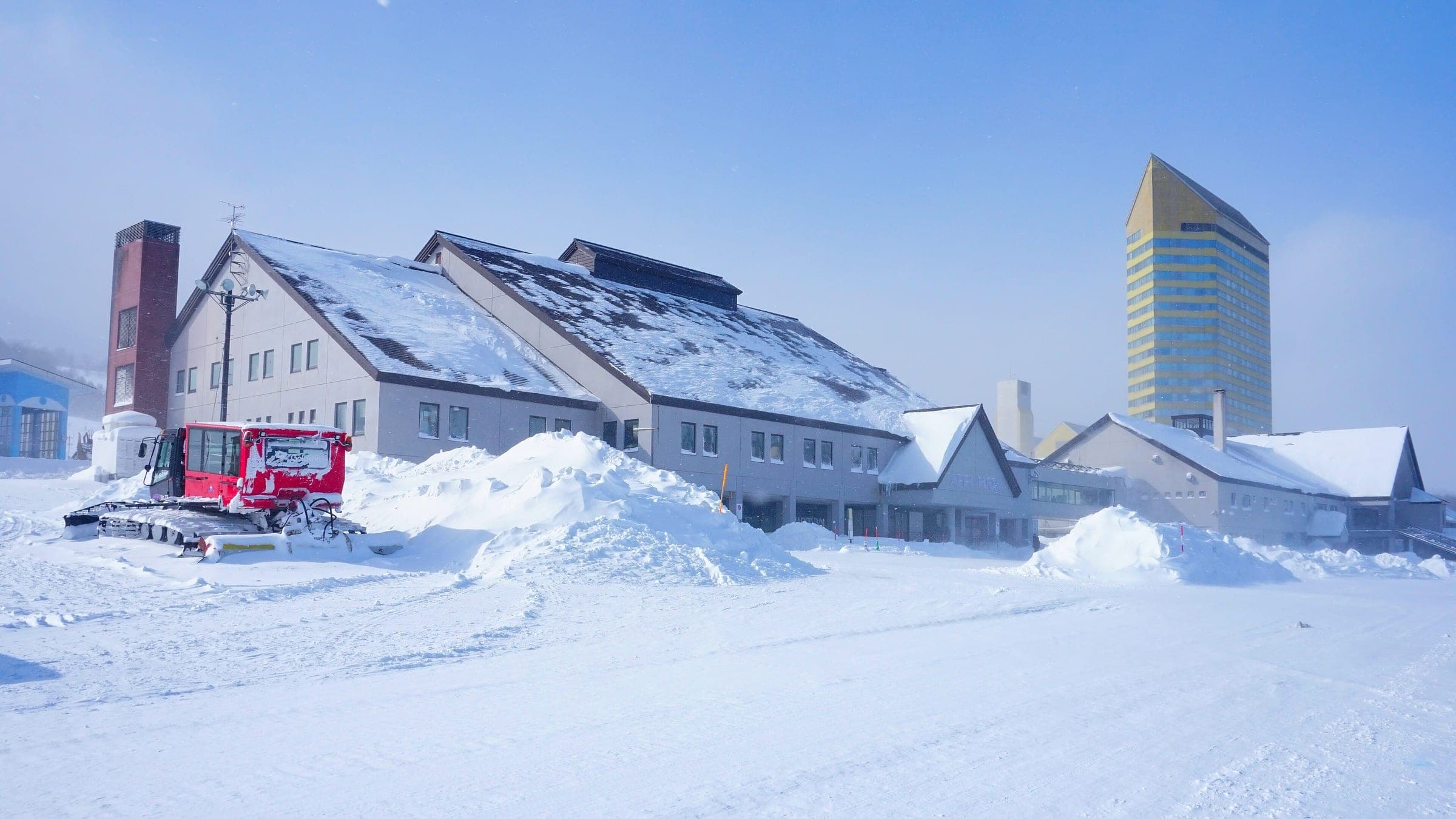 A snowcat parks beside the snowed-in base lodges at APPI with the hotel tower behind.
