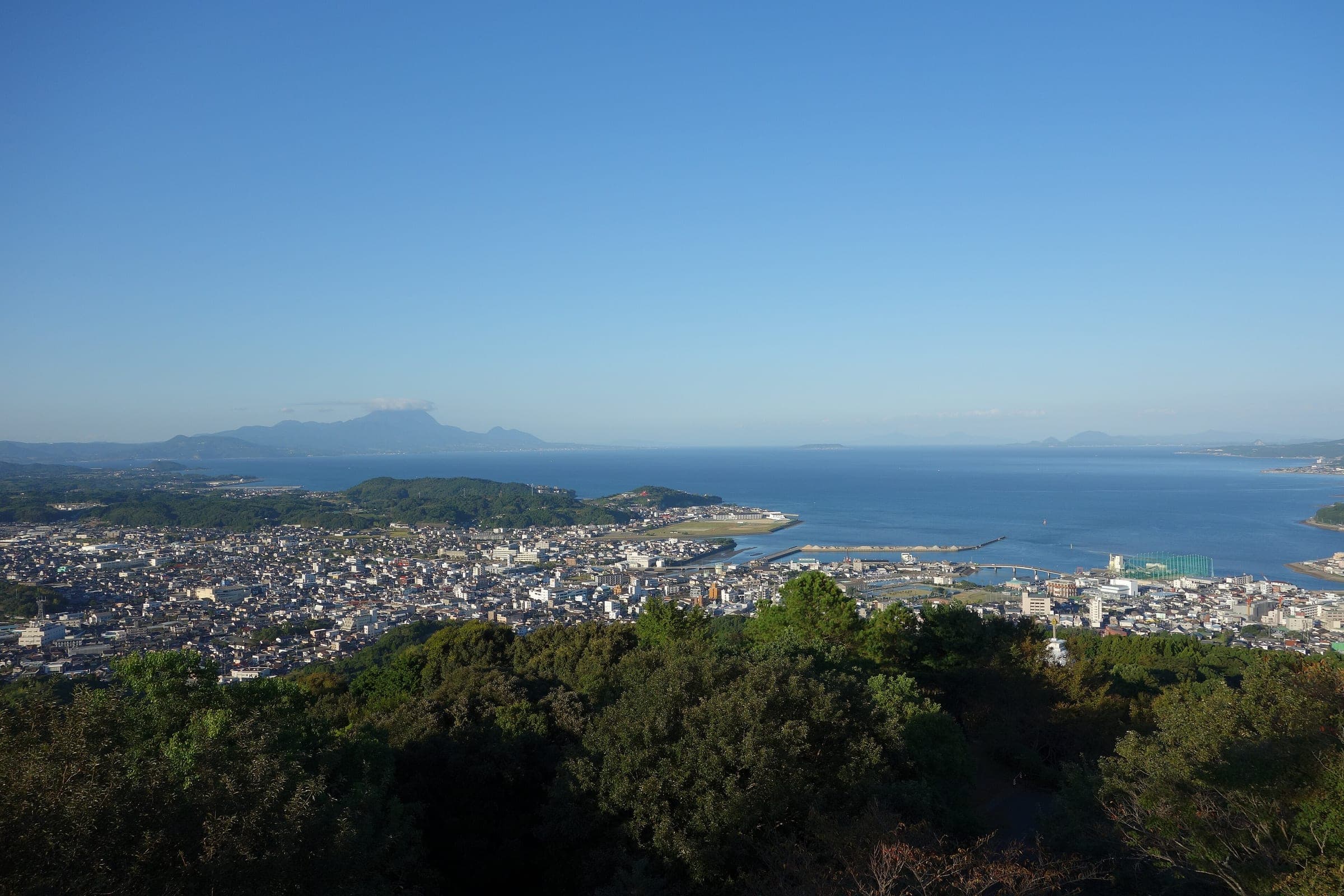 Hondo's rooftops and harbour spread along the shore below Jumanyama with the sea and distant peaks beyond.